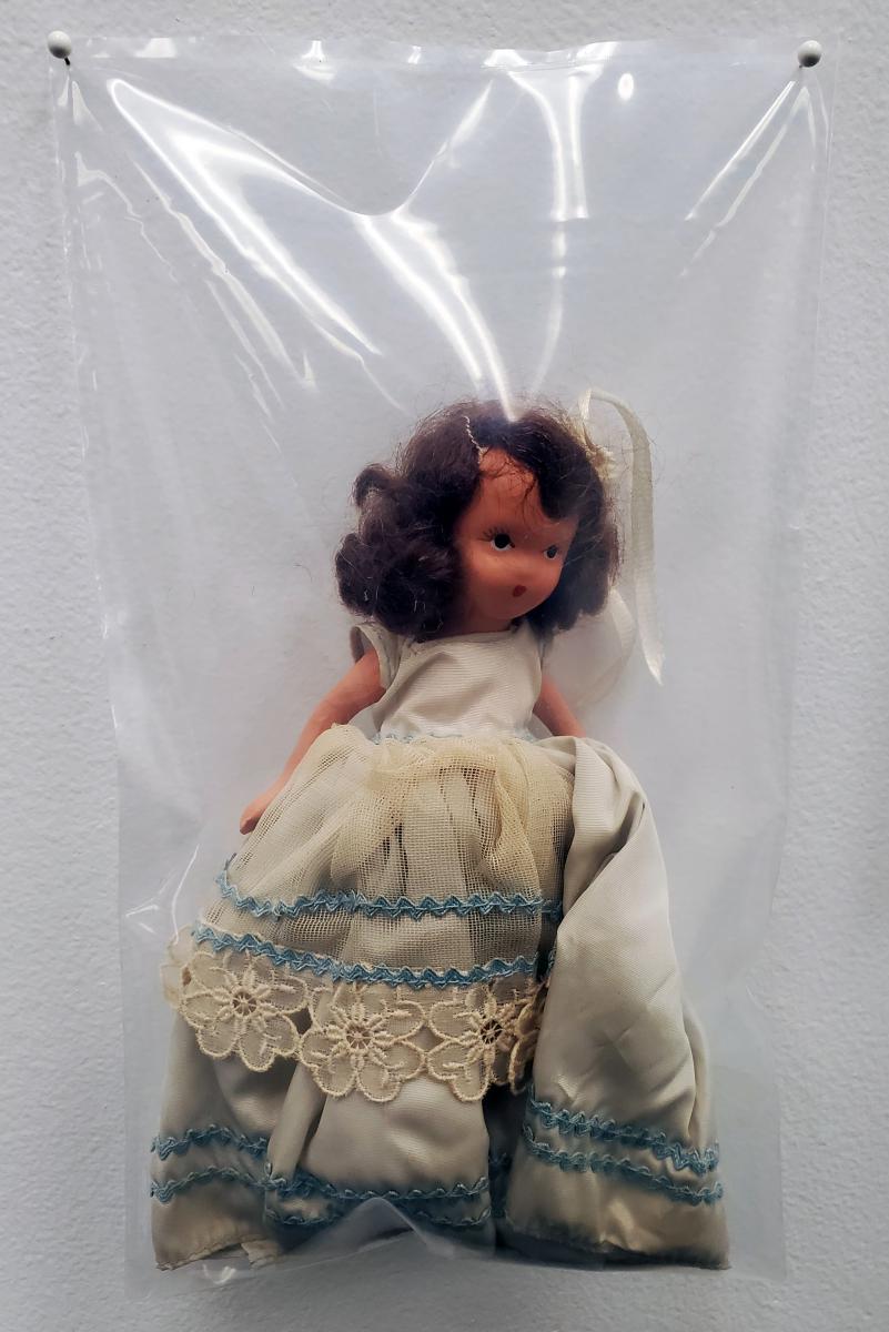 A white doll with brown hair wearing a cream dress is in a clear plastic bag tht is pinned to the wall.