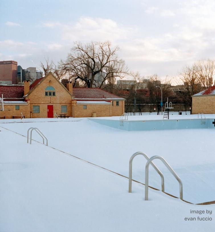 Color image of an empty swimming pool filled with snow, buildings and bare trees rise on the skyline